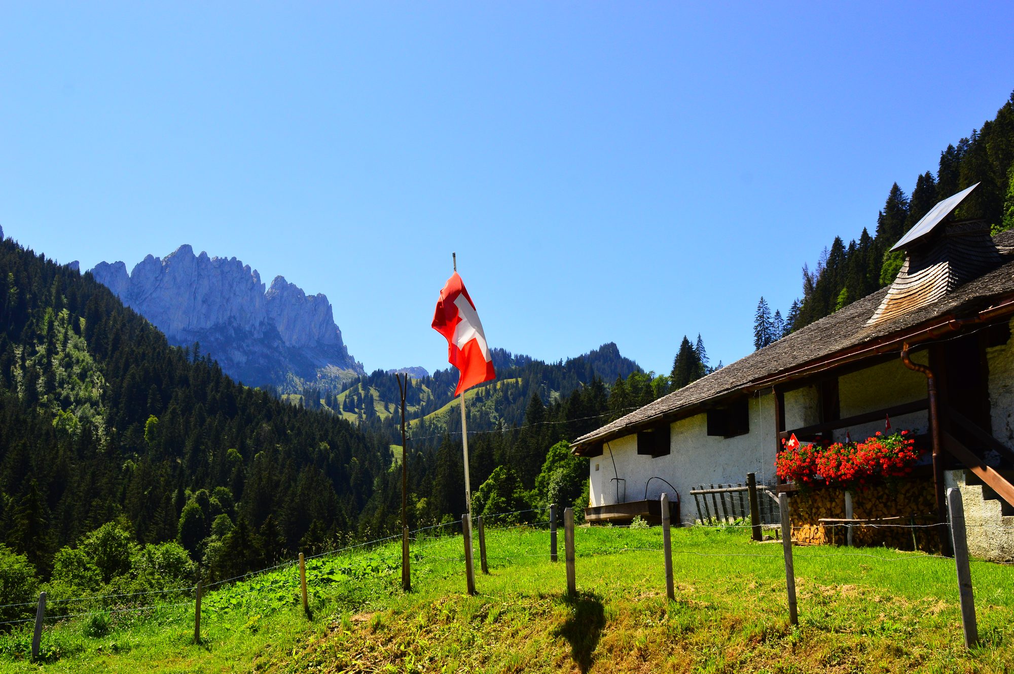 A Swiss flag flying beside an alpine chalet with red geraniums, the Gastlosen behind